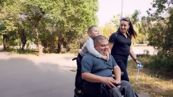 車椅子の男。ハンディキャップの男。自動運転車椅子の若い障害者は家族や妻や小さな子供と一緒に晴れた秋の日に街の路地を歩きます. — ストック動画