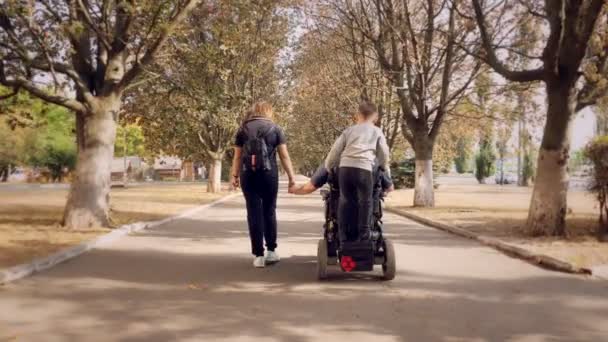 車椅子の男。ハンディキャップの男。自動運転車椅子の若い障害者は晴れた秋の日に家族と妻と小さな子供と街の路地を歩きます。バックビュー — ストック動画