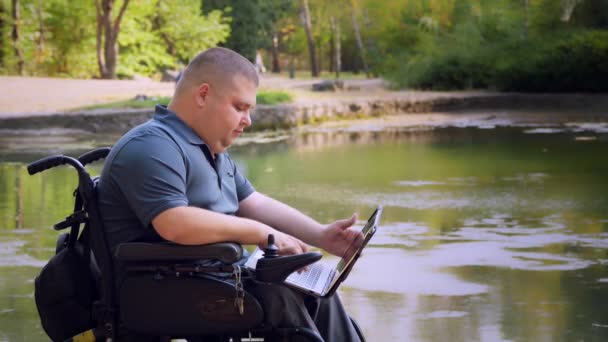 Wheelchair man. Handicapped man. young disabled man, sitting in an automated wheelchair and working on a laptop, in a city park, by the lake, on autumn sunny day. — Stock Video