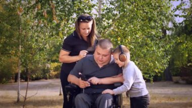 Tekerlekli sandalye. Engelli adam. Genç engelli bir adam tekerlekli sandalyede oturuyor, etrafı ailesi, karısı ve küçük oğluyla çevrili. Müzik dinlerler, akıllı telefondan video izlerler, rahatlarlar. Şehir parkında, sonbaharda.