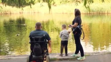 Tekerlekli sandalye. Engelli adam. Genç engelli bir adam tekerlekli sandalyede oturuyor, etrafı ailesi, karısı ve küçük oğluyla çevrili. Göldeki ördekleri besler, güneşli bir sonbahar gününde şehir parkında rahatlarlar.