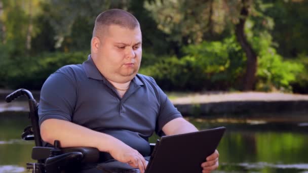Инвалид-колясочник. Инвалид. молодой инвалид, сидящий в автоматическом инвалидном кресле и работающий на ноутбуке, в городском парке, у озера, осенью солнечный день. — стоковое видео