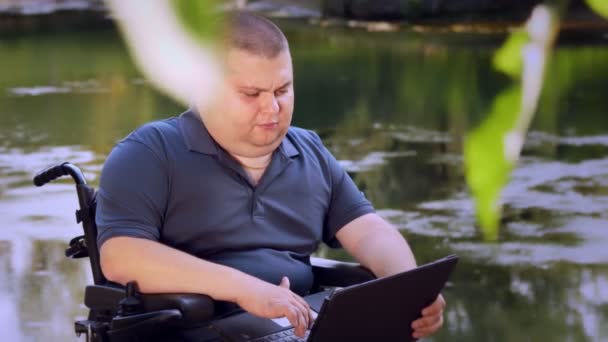 Kerekesszékes. Fogyatékos ember. fiatal fogyatékos férfi, ül egy automatizált kerekesszékben, és dolgozik egy laptop, egy városi parkban, a tó mellett, ősszel napsütéses napon. — Stock videók