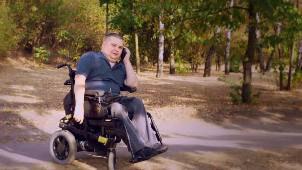 Wheelchair man. Handicapped man. young disabled man sitting in an automated wheelchair and talking on a cell phone, in a city park, on an autumn sunny day. — Stock Video