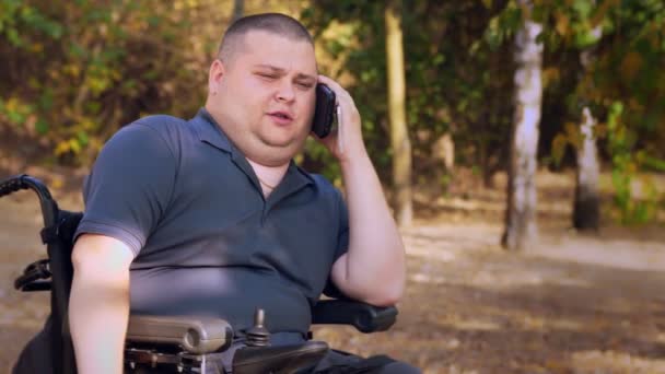 Homem de cadeira de rodas. Homem deficiente. jovem deficiente sentado em uma cadeira de rodas automatizada e falando em um telefone celular, em um parque da cidade, em um dia ensolarado outono. — Vídeo de Stock