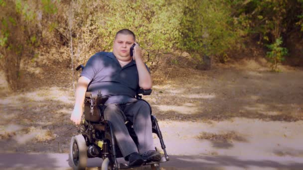 Wheelchair man. Handicapped man. young disabled man sitting in an automated wheelchair and talking on a cell phone, in a city park, on an autumn sunny day. — Stock Video