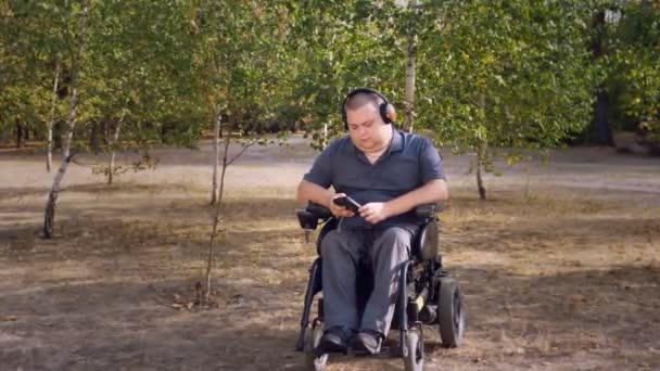 Wheelchair man. Handicapped man. young disabled man, sitting in an automated wheelchair and listening to music on headphones, in a city park, on an autumn sunny day. — Stock Video