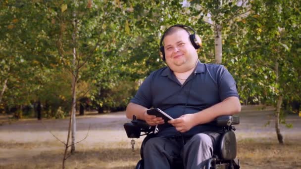 Wheelchair man. Handicapped man. young disabled man, sitting in an automated wheelchair and listening to music on headphones, in a city park, on an autumn sunny day. — Stock Video