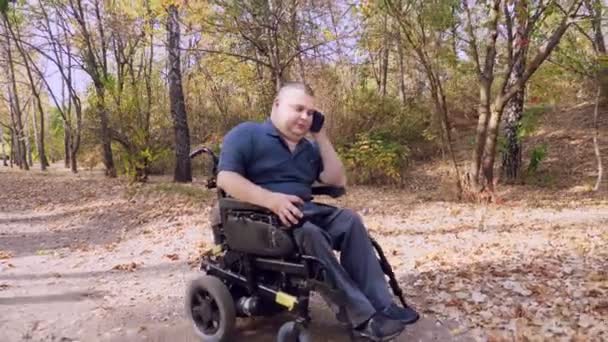 Wheelchair man. Handicapped man. young disabled man rides in an automated wheelchair and talking on a cell phone, in a city park, on an autumn sunny day. — Stock Video