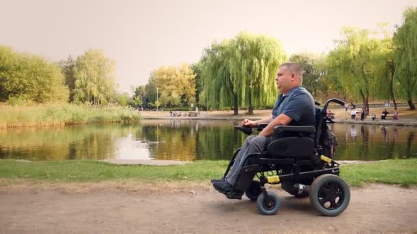 Homme en fauteuil roulant. Homme handicapé. un jeune handicapé dans un fauteuil roulant automatique marche au bord du lac, dans un parc municipal, par une journée ensoleillée d'automne. — Video