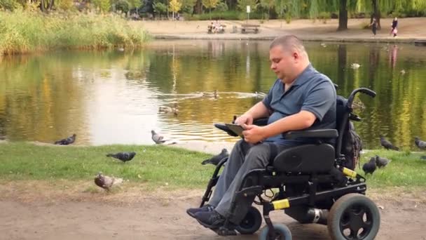Wheelchair man. Handicapped man. young disabled man, sitting in an automated wheelchair and working on digital tablet, in city park, by the lake, on autumn sunny day. — Stock Video