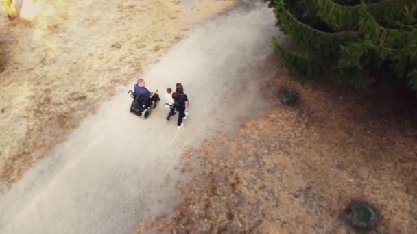 Tekerlekli sandalye. Engelli adam. Otomatik tekerlekli sandalyedeki genç adam güneşli bir sonbahar gününde ailesi, karısı ve küçük çocuğuyla parkta yürüyor. Arkadan bak. Aero — Stok video