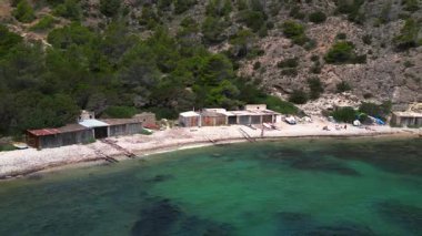 İspanya 'nın İbiza eyaletinin Cala Llentrisca kentindeki sığ sulara giden bir tahta merdiveni olan çakıl taşlı sahildeki eski kayıkhanelerin önündeki kaplumbağa. Geri geri uç. Çok yakın. Uçağı geç. 