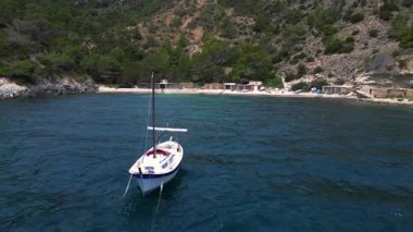Ibiza 'da bazı balıkçıların evlerinin olduğu gizli bir sahil koyunun yakınında sakin mavi sularda yüzen yelkenli. Ters İHA uçuşu 