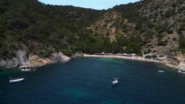 Ibiza 'da bazı balıkçıların evlerinin olduğu gizli bir sahil koyunun yakınında sakin mavi sularda yüzen yelkenli. İHA alçalıyor 