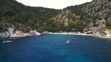 Ibiza 'da bazı balıkçıların evlerinin olduğu gizli bir sahil koyunun yakınında sakin mavi sularda yüzen yelkenli. panorama genel görünüm dronu 