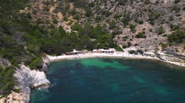 Yazın İspanya 'nın İbiza kentindeki güzel Cala Llentrisca sahilinin turkuaz suyu ve Barques de Pescadors Huts manzarası. panorama yörünge dronu 