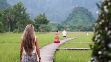 Laos kırsalında yemyeşil bir çeltik tarlasında tahta bir kaldırımda yürüyen bir kadının arka görüntüsü.
