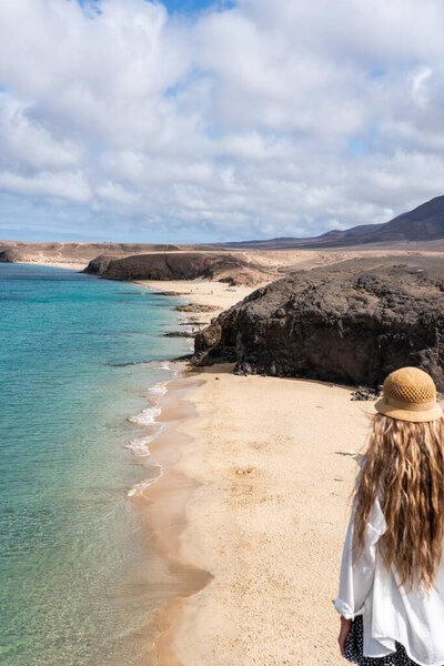 A woman with long hair in a sun hat stands on a sandy Papagallo beach, gazing at the turquoise water and rocky coastline in Lanzarote