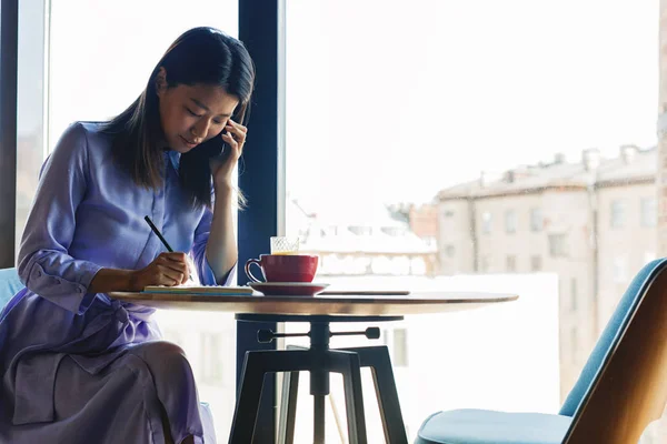 Asian woman talking on mobile phone. Young woman sitting in cafe and writing on notepad.