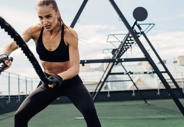 Woman using battle ropes during strength training on rooftop. Young female working out outdoors.