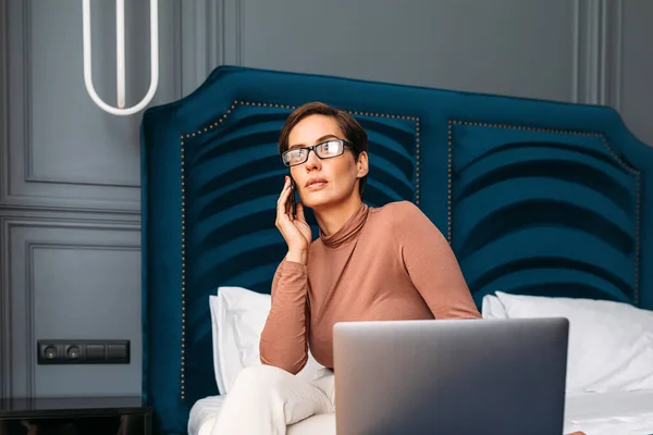 Caucasian woman in casual clothes sitting on bed in hotel room using smartphone and looking at window. Businesswoman working from hotel room.