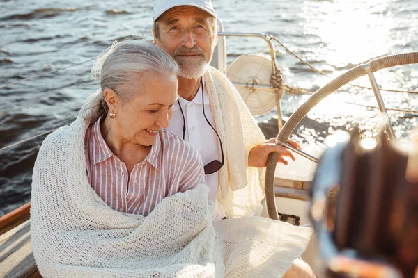 A couple of two mature people wrapped in plaid on a sailboat. Senior man in cap steering a yacht.