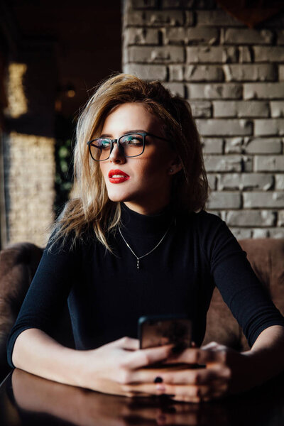 Young smiling business woman sitting in cafe at table, leaning hand on table and holding smartphone.