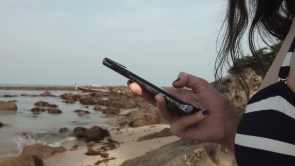 Ragazza utilizza un telefono cellulare sulla spiaggia sullo sfondo del mare e pietre. scrive un messaggio . — Video Stock