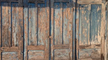 Bir çift yıpranmış ahşap kapılı, mavi boyalı ve görünür, aşınmış ahşap. Soyulmuş boyanın dokusu ve doğal ahşap tanecikleri yüzeye vuran sıcak güneş ışığıyla vurgulanır..