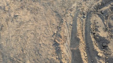 Değişik dokuları olan kuru, kahverengi bir dünya. Sağ tarafta derin bir lastik izi ve dağılmış kayalar var. Güçlü gölgeler oluşturuyor. Sol tarafta ise daha pürüzsüz bir toprak ve soluk izler var..