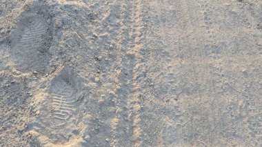 Açık renkli, tozlu toprağın yüksek açılı görüntüsü. Dokulu zemin iki farklı ayakkabı izi ve belli belirsiz bir lastik izi ile işaretlenmiş. Bu da zamanın ve insan aktivitesinin geçişini gösteriyor..