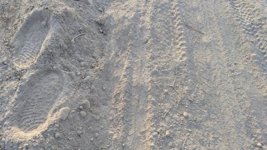 Açık renkli, tozlu toprağın ve ince çakılların yukarıdan görünüşü. Zemin, pürüzsüz bir ayakkabı izi ve zayıf bir lastik izi ile işaretlenmiş, engebeli arazide insan ve makine izlerinin bir karışımını yakalıyor..