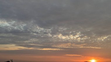 Karanlık bulutlardan bir battaniyeyle gün doğumu gökyüzü. Güneş sadece ufukta görünür. Canlı turuncu ve sarı bir parıltı yayar. Bulutların altını aydınlatır ve dramatik bir sahne yaratır..