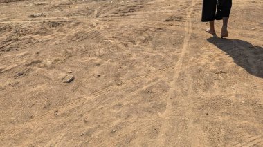 Bir insanın ayakları tozlu toprak bir yolda yürür. Zemin lastik izleri ve ince kumlu toprakla kaplı. Kişinin uzun bir gölgesi dokulu yüzeyine düşer..