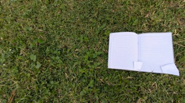 Yemyeşil çimenlerin üzerinde, üzerinde kurallı sayfalar bulunan açık bir defter var. Resim yukarıdan aşağıya doğru bir perspektiften çekilmiş, defter çerçevenin sağ tarafına yerleştirilmiş..