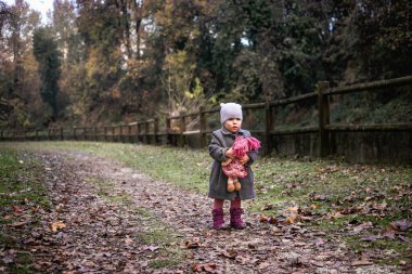 Gri montlu küçük kız elinde pembe saçlı yumuşak tekstil bebeği açık havada ahşap çitin yanında duruyor.