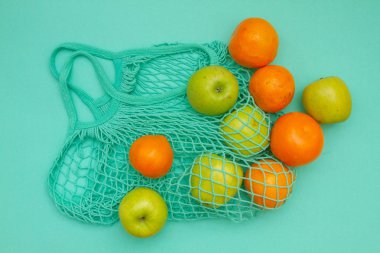 Örülmüş bir nane rengi ip torbasında meyveler vardır. Mandalinalar, portakallar ve elmalar açık mavi bir arkaplanda bir alışveriş torbasında.