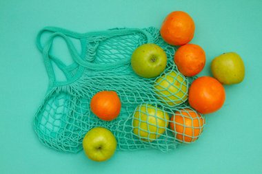 Örülmüş bir nane rengi ip torbasında meyveler vardır. Mandalinalar, portakallar ve elmalar açık mavi bir arkaplanda bir alışveriş torbasında.