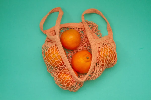 Tangerines in green mint color string bag on a turquoise background. Citrus in a wicker bag on paper. Shot from above.