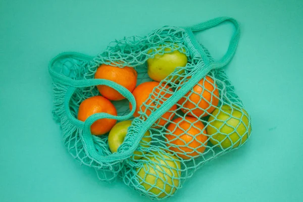In a woven neo mint color string bag there are fruits. Tangerines and oranges and apples in a shopping bag on a light blue mint background.