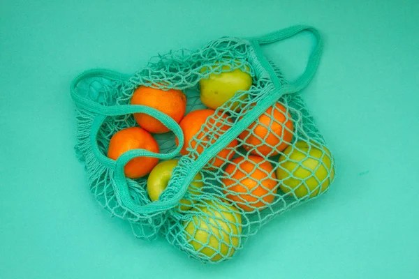 In a woven neo mint color string bag there are fruits. Tangerines and oranges and apples in a shopping bag on a light blue mint background.