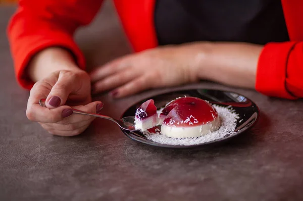 Woman\'s hands in a red jacket. The girl is eating cake. Hemisphere red jelly on a black plate. Sweetness is sprinkled with coconut