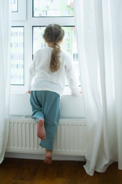 Parlak bir odada küçük şirin bir kız, odada en az sayıda eşya, çocuk seviniyor, beyaz perdelerle pencerenin kenarında oturuyor..