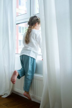 Parlak bir odada küçük şirin bir kız, odada en az sayıda eşya, çocuk seviniyor, beyaz perdelerle pencerenin kenarında oturuyor..