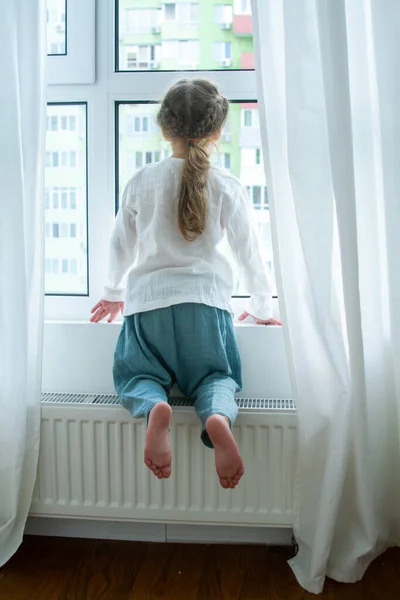 Parlak bir odada küçük şirin bir kız, odada en az sayıda eşya, çocuk seviniyor, beyaz perdelerle pencerenin kenarında oturuyor..