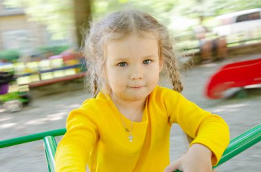 Sarı tişörtlü güzel bir kız oyun parkında oynuyor, çocuk atlıkarıncaya biniyor, hareket hissi uyandırıyor..