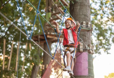 Turuncu miğferli ve özel korumalı küçük bir kız halat parkında engellere takılıyor. Çocukların boş zamanı, çocuklar için bir oyun alanı, parkta eğlence..