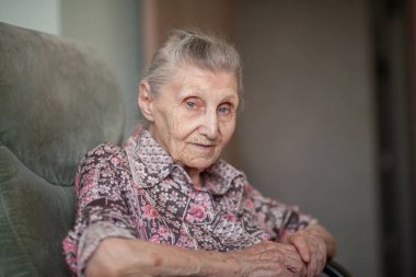 Pembe bluzlu yaşlı bir kadın evde bir sandalyede oturuyor, bir büyükanne gülümsüyor, kadınlarda yaşlılık..