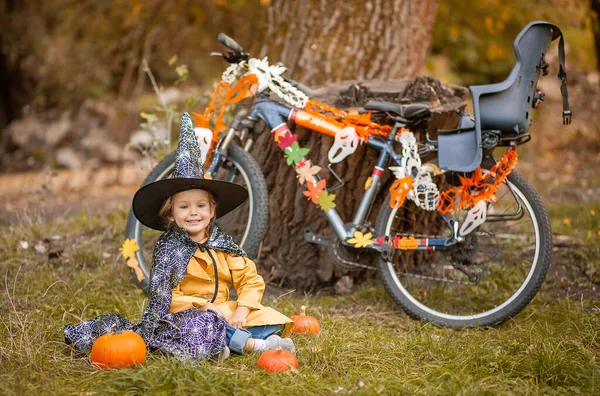 自然の中でハロウィーンの休日に小さな女の子 子供はガーランドで飾られた自転車の近くに座っています — ストック写真